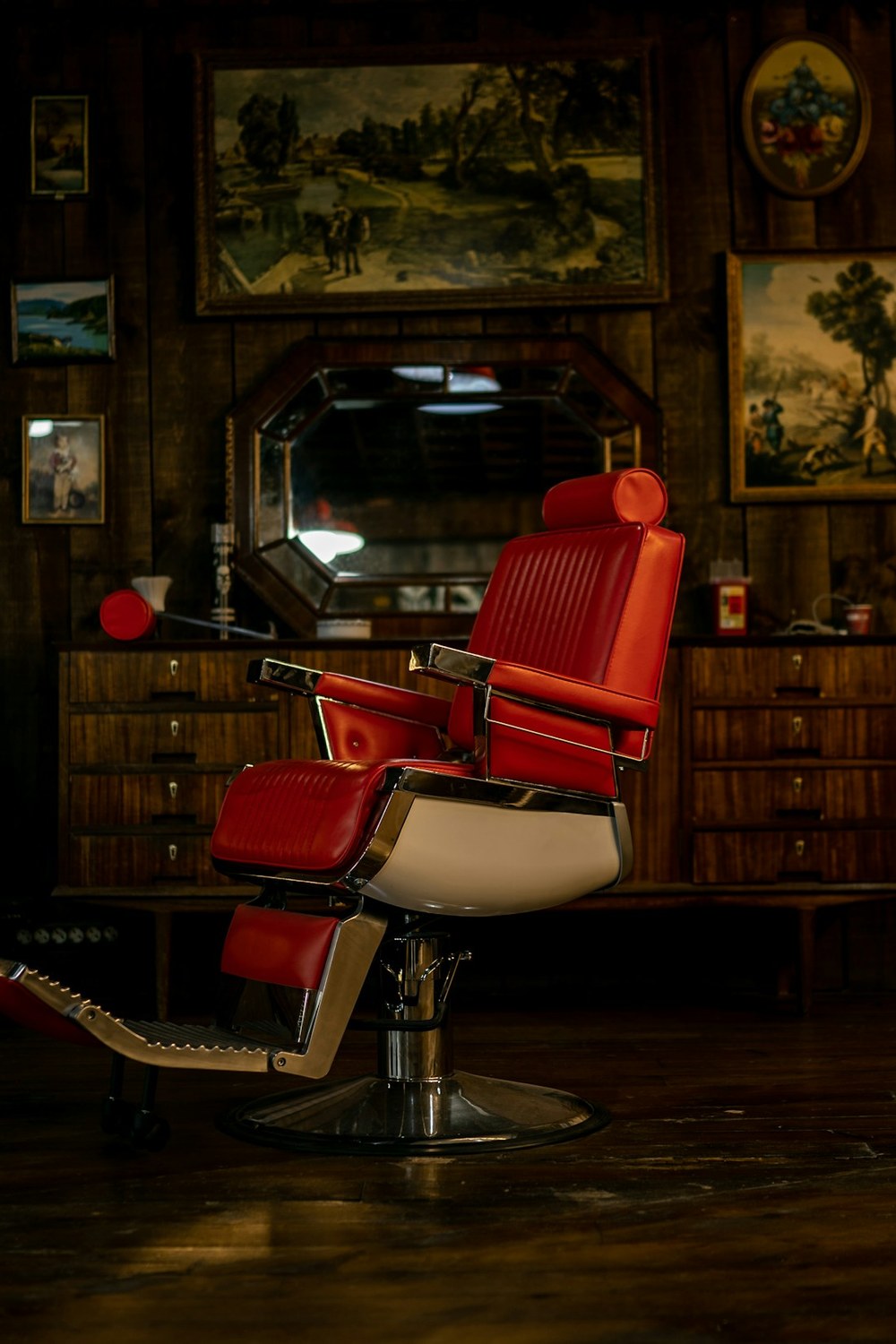 A red leather barber chair in the middle of a warm, wood-panelled room hung with framed paintings, lit by one lamp
